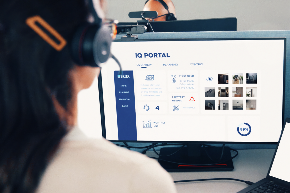A person wearing a headset sits at a desk in a modern office cubicle, facing a computer monitor displaying the BRITA iQ Portal, with a laptop in front and a second monitor behind them.