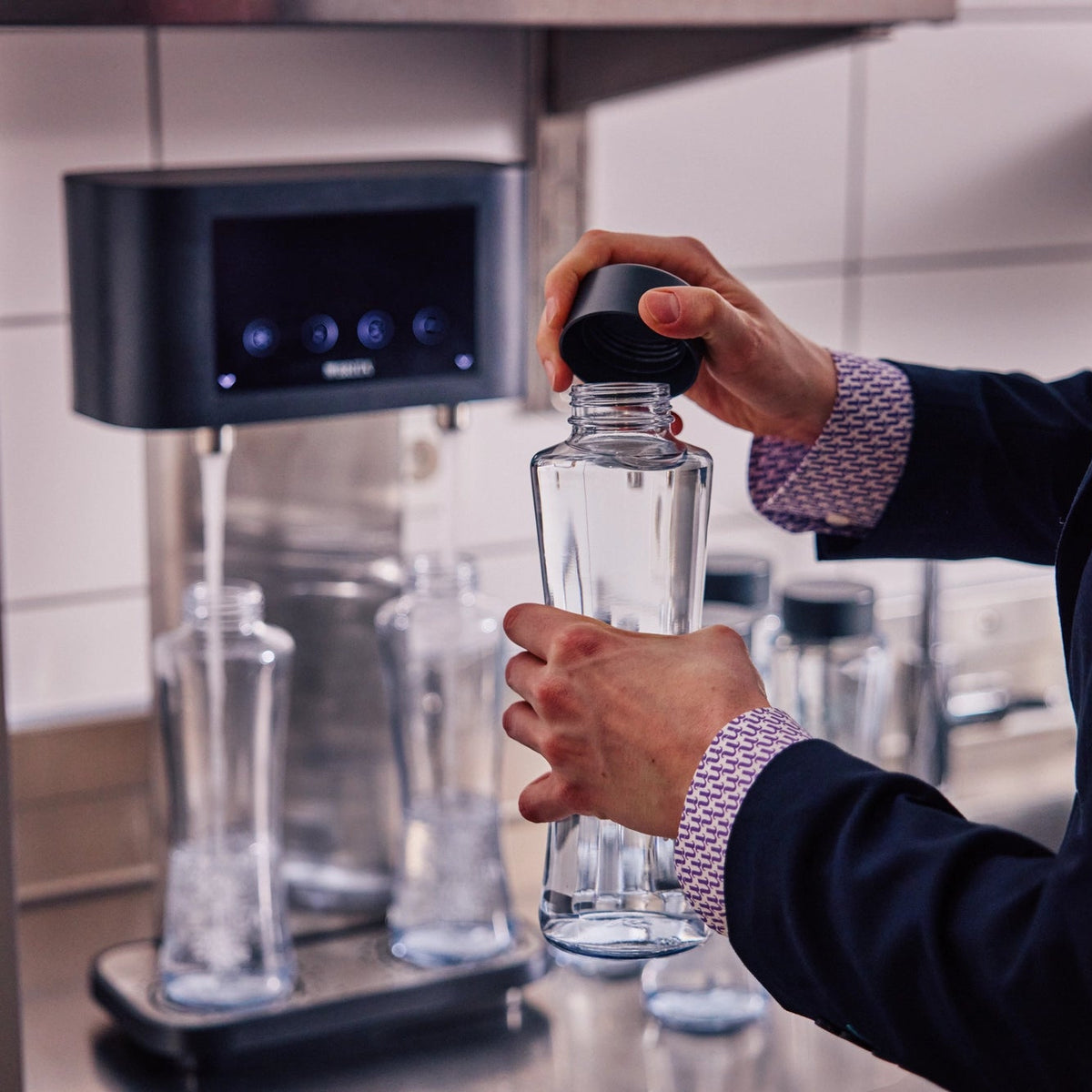 Person holding a BRITA Twist bottle with a dark cap in front of a BRITA Fill T-Tap.