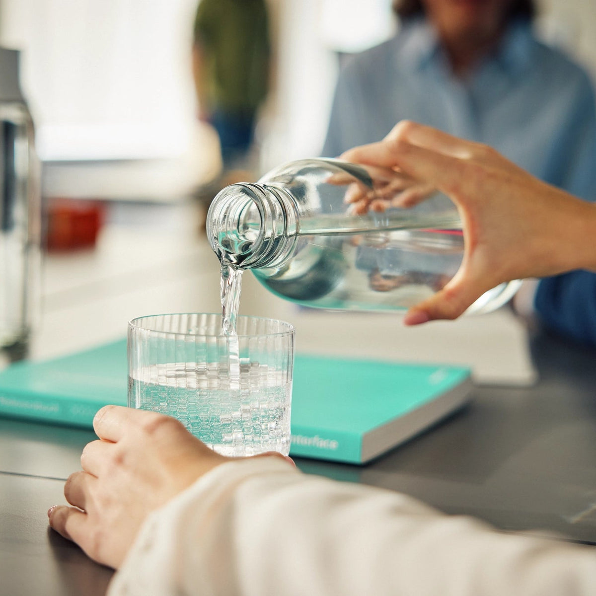 A person pouring water from a bottle into a glass which is standing on an office table.